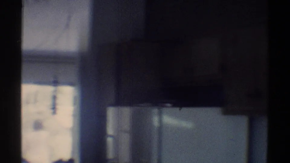 Soft 8mm frame of the kitchen pan showing dark cabinetry, a white refrigerator, and daylight entering from a window or doorway.