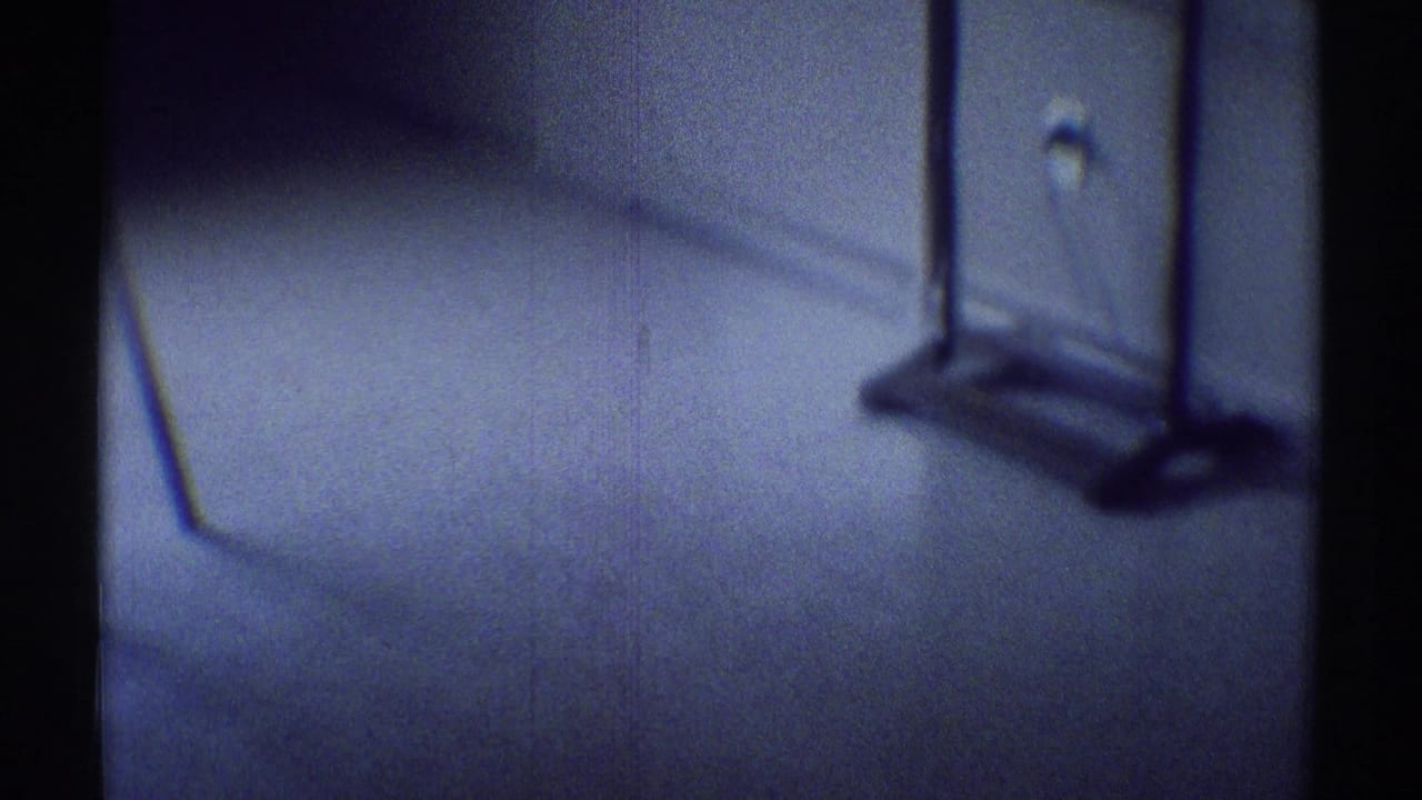 1969 8mm frame from a dark Woodland Hills house interior showing carpet, wall trim, outlet, and a coat rack or stand in soft focus.