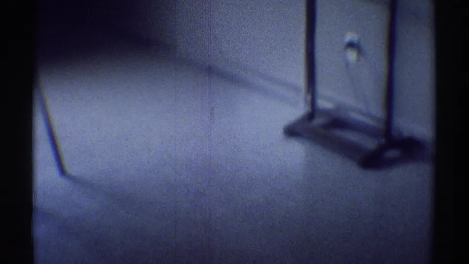 1969 8mm frame from a dark Woodland Hills house interior showing carpet, wall trim, outlet, and a coat rack or stand in soft focus.