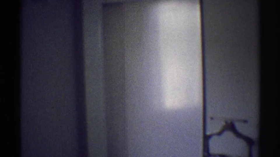 Low-light 8mm frame inside a Woodland Hills house, showing a dark wall, carpeted floor, and partial coat rack or stand near the lower edge.