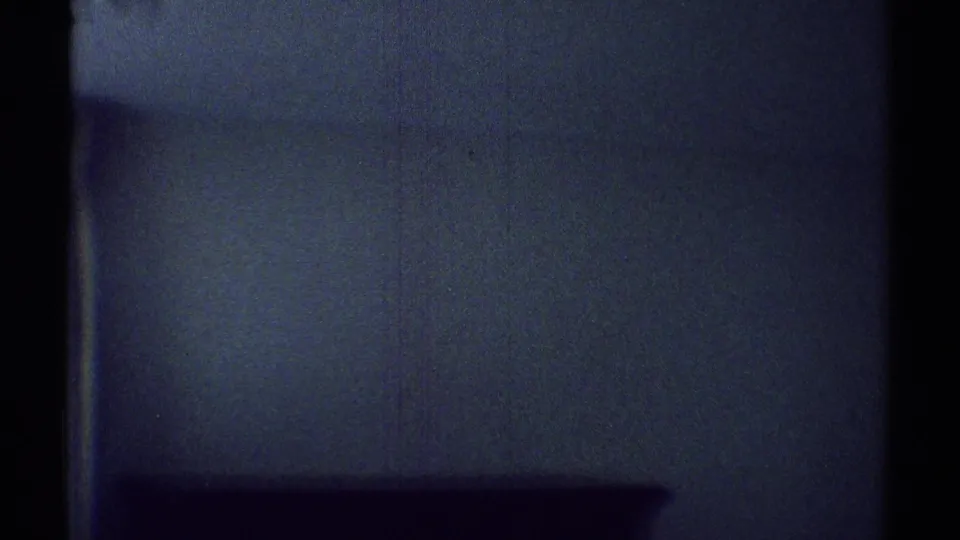 1969 Woodland Hills home-movie frame showing a darkened room wall, faint curtain edge, and low shadowed interior shape.
