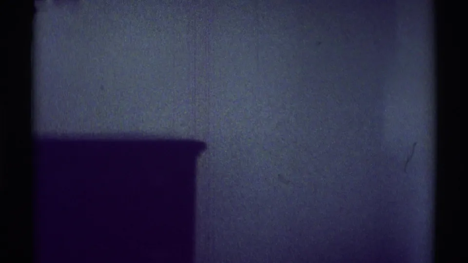 Dim 1969 Woodland Hills bedroom view with a dark furniture silhouette against a textured wall on 8mm film.
