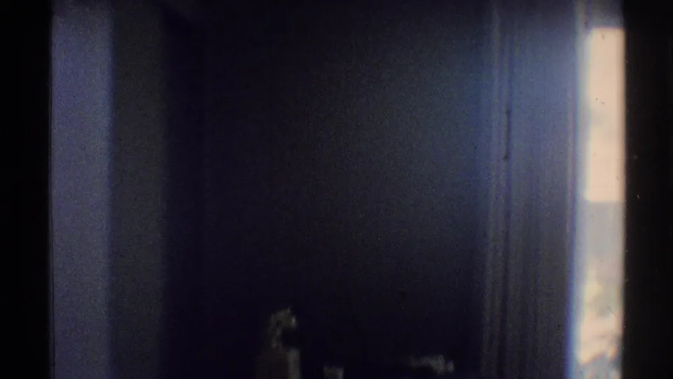 Dim Woodland Hills bathroom frame showing a dark wall, small fixture area, and bright window edge in grainy 8mm light.