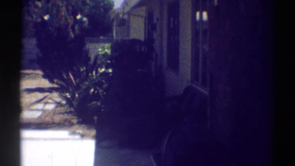 Woodland Hills 1969 home-movie frame showing a backyard path, house wall, shrubs, and heavy shadow along the right side.