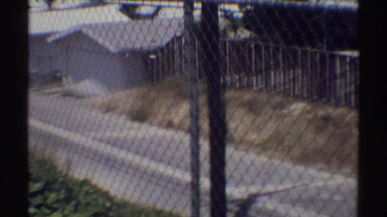 1969 Woodland Hills residential view through a chain-link fence, with a paved street and modest single-story homes beyond.