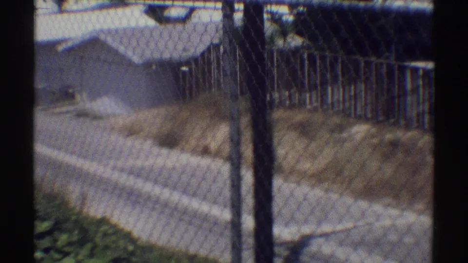 1969 Woodland Hills residential view through a chain-link fence, with a paved street and modest single-story homes beyond.