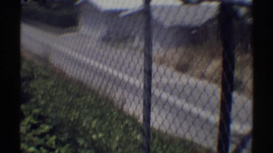 Woodland Hills home-movie frame showing a fence line, grass, and a quiet residential street in soft 8mm color.