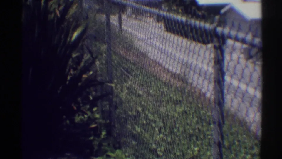 Handheld 8mm view along a chain-link fence, with greenery, a sidewalk edge, and suburban houses beyond.