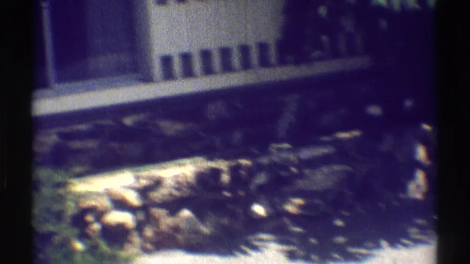 1969 Woodland Hills home-movie frame showing a white house exterior, dark windows, stone wall, and sunlit plants in the yard.