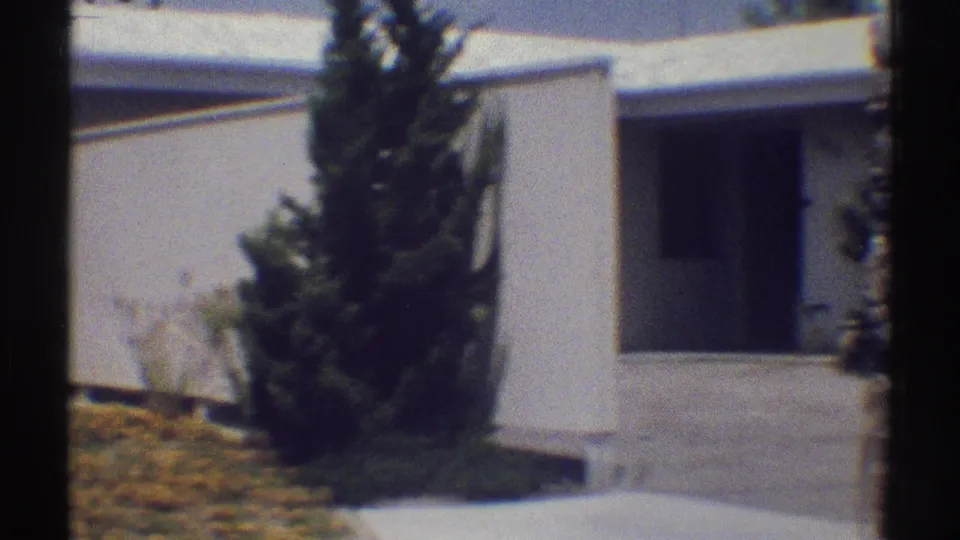 1969 Woodland Hills frame showing a white house entrance, tall evergreen, low plantings, and sunlit walkway.