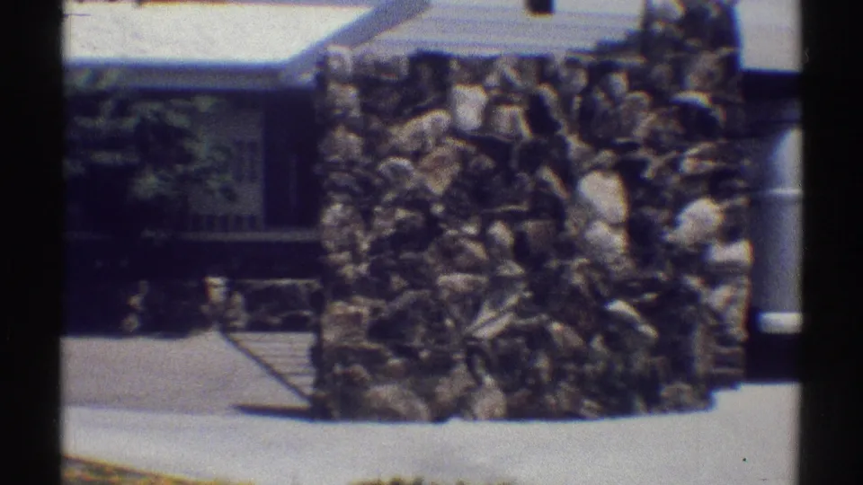 Rock masonry feature, driveway pavement, and part of a white Woodland Hills home exterior in a 1969 8mm frame.