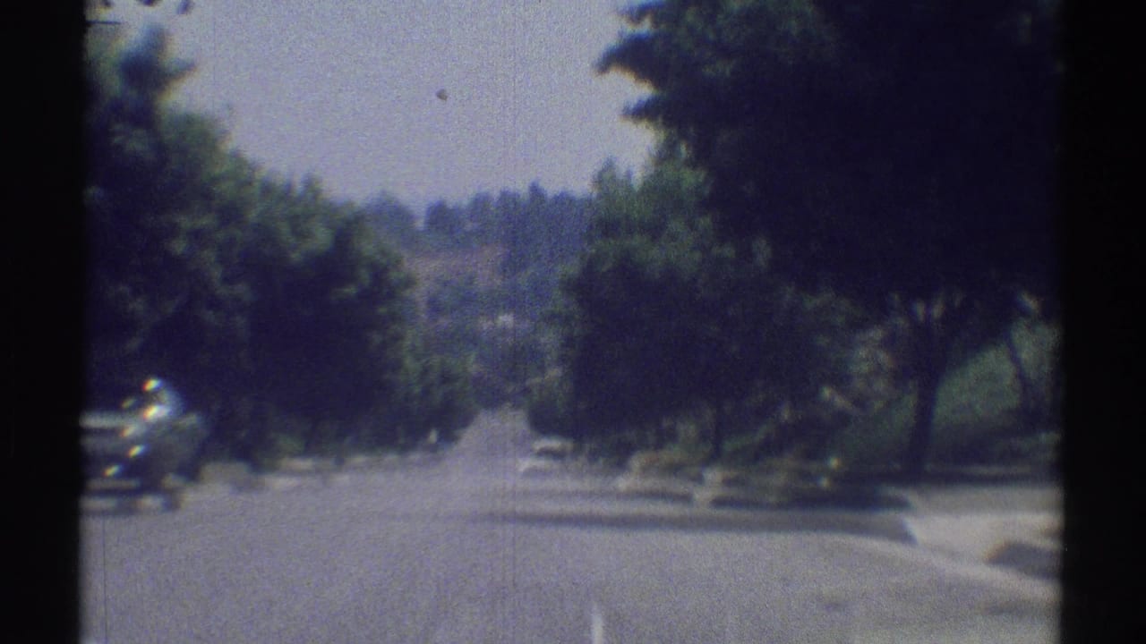 1969 8mm view of a Woodland Hills residential street from a moving vehicle, with trees, curb, parked car, and hills in the distance.