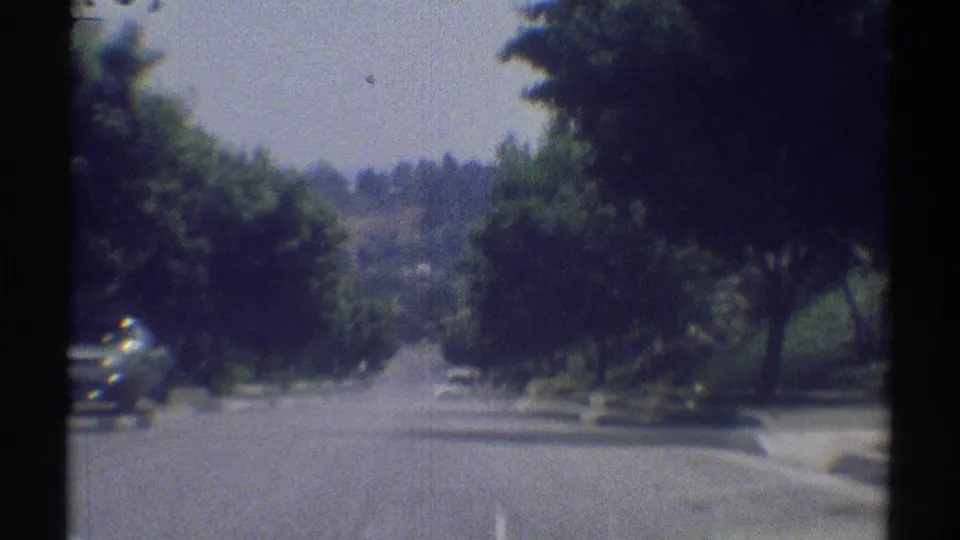 1969 8mm view of a Woodland Hills residential street from a moving vehicle, with trees, curb, parked car, and hills in the distance.