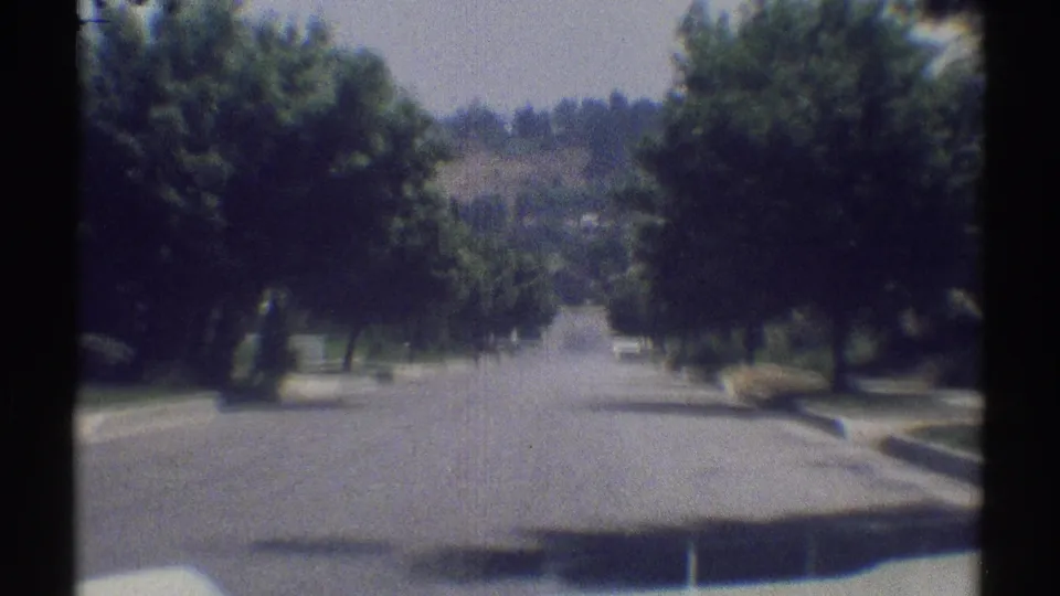 Woodland Hills street view from 1969 home-movie footage, showing a broad road, leafy trees, lawns, and distant hillside houses.