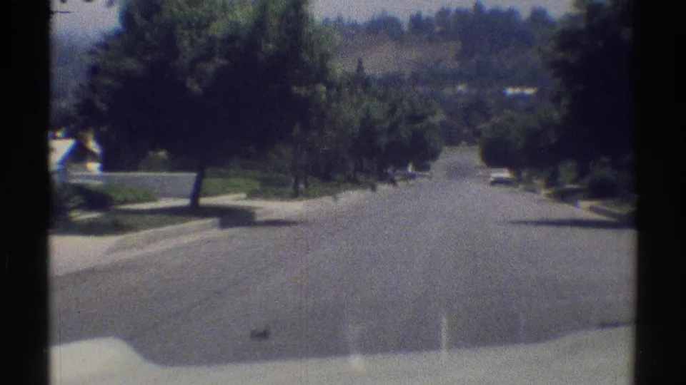 End frame of a 1969 Woodland Hills drive, looking down a sunlit residential street toward distant hills and a small car ahead.