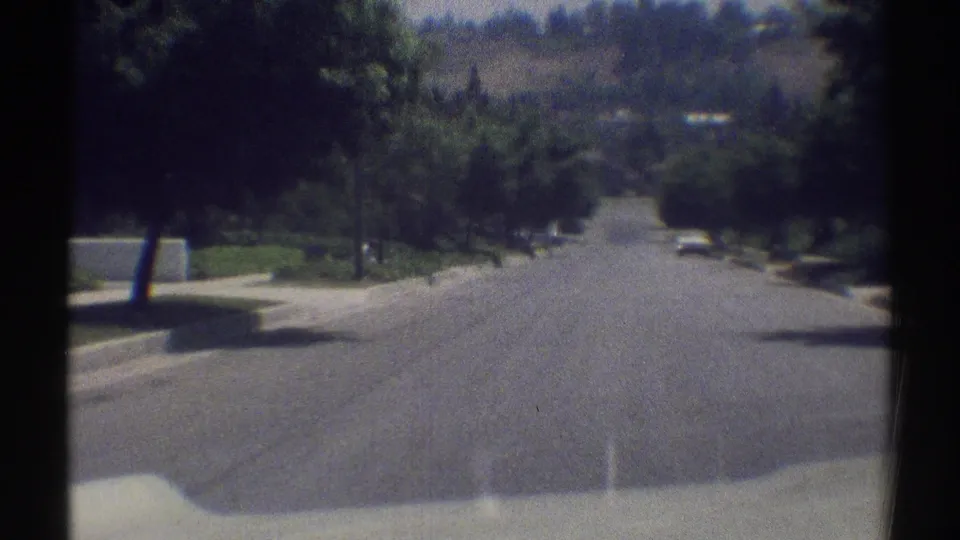 1969 Woodland Hills home-movie frame looking forward along a broad suburban street with trees, sidewalks, and a small white car ahead.