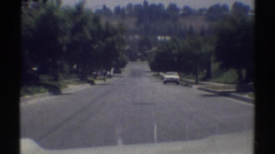 1969 Woodland Hills frame from a moving vehicle, showing a sunny residential road and a white car parked along the right curb.