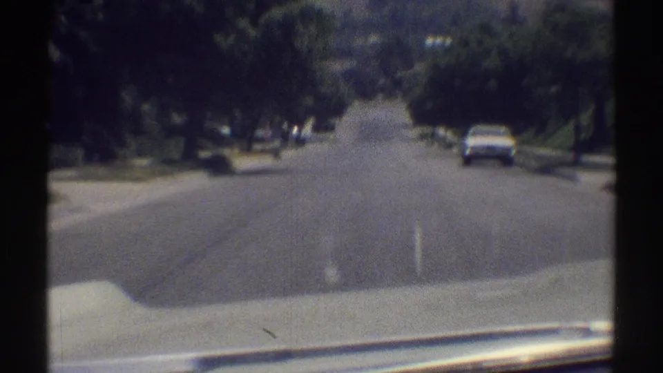1969 Woodland Hills home-movie view nearing a white parked car on a quiet suburban street bordered by trees and lawns.