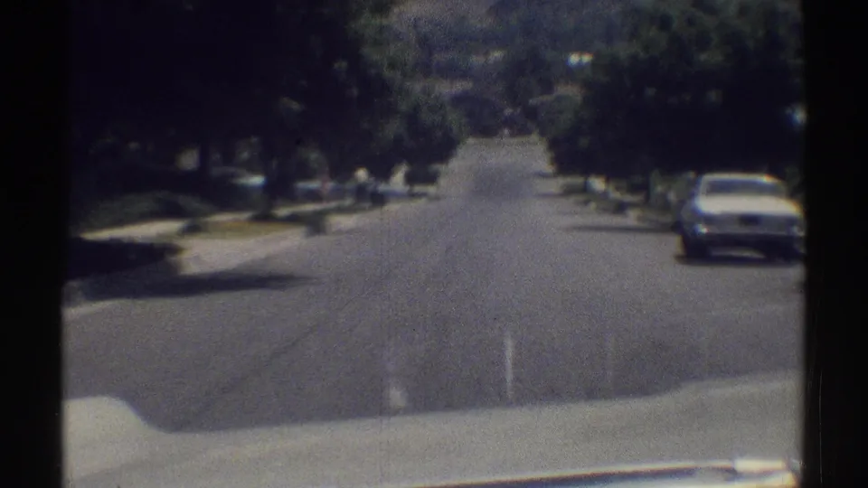Final 1969 Woodland Hills frame showing the moving view passing closer to a white car parked along the right curb of a tree-lined street.