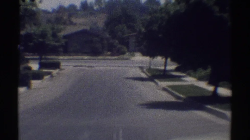 1969 Woodland Hills street view from a moving vehicle, showing a quiet residential road, curb lawns, trees, and low houses ahead.