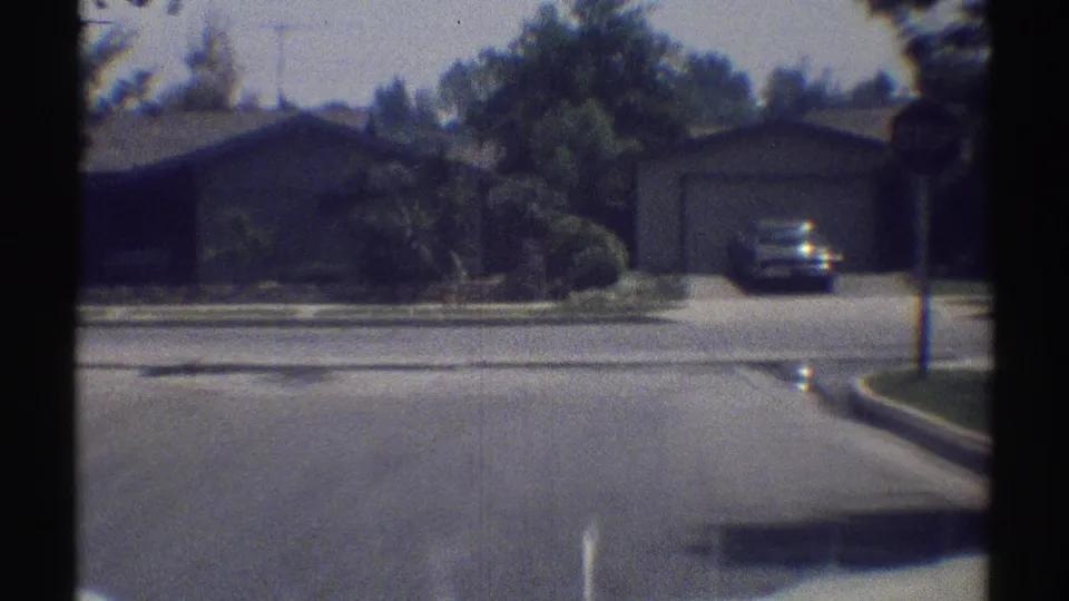 1969 Woodland Hills home-movie frame showing a single-story house, driveway, dark sedan, curb, and sunlit residential street.
