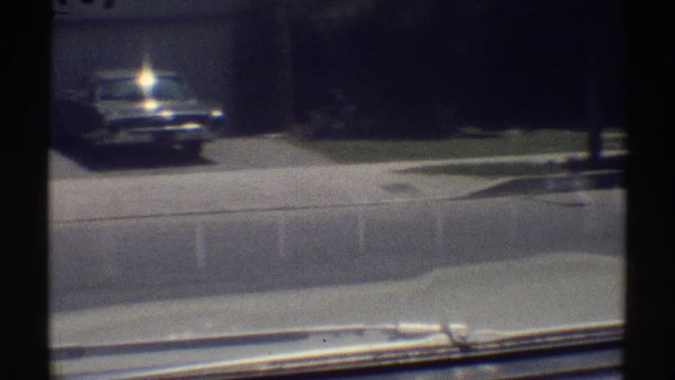 1969 Woodland Hills vehicle-perspective frame with a parked sedan, curb, lawn, shade trees, and part of the filming vehicle visible.