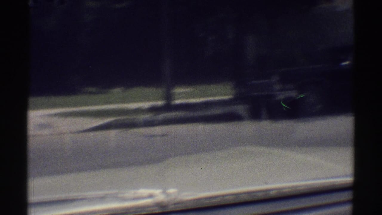 View through a car windshield onto a quiet suburban street with trees and a dark vehicle ahead.