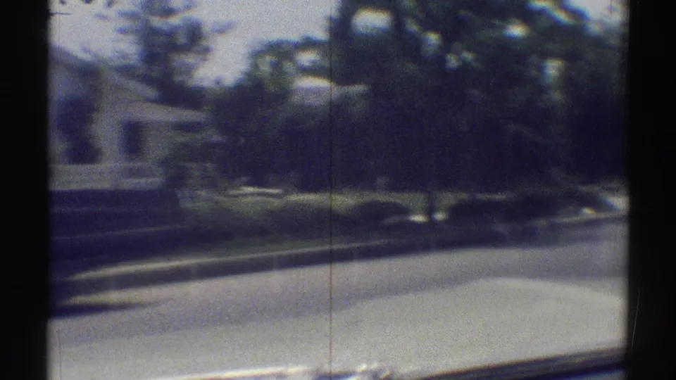 1969 Woodland Hills street view from a moving car, with a low residential building, trees, lawn, and curved roadway visible through soft 8mm focus.