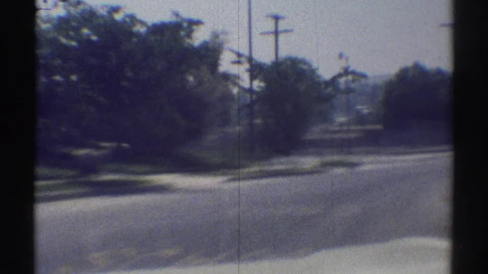 Soft 8mm view of a Woodland Hills residential street in 1969, filmed from a moving car with trees, utility poles, and bright roadway ahead.