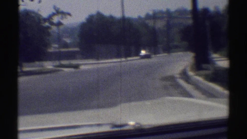 1969 Woodland Hills car-ride frame showing a curved suburban street, trees, curb, car hood edge, and a low residential building beyond.