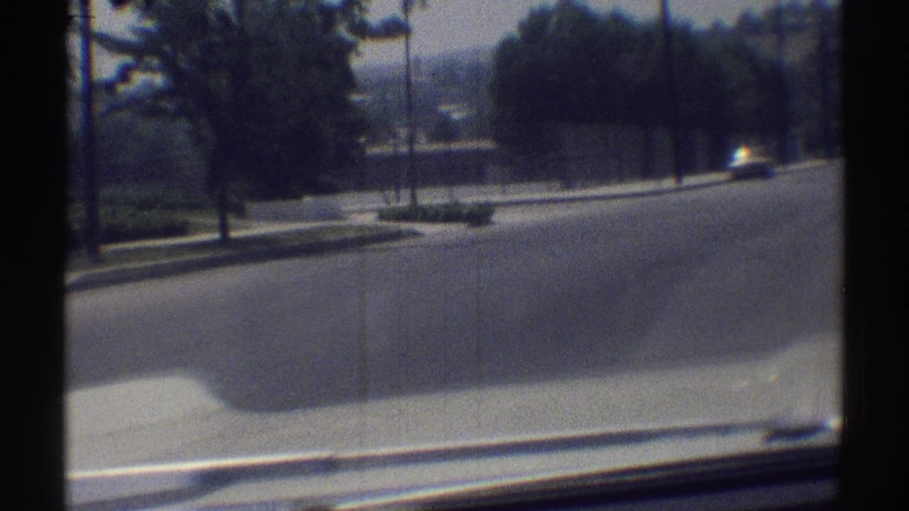 View from a moving car past a quiet street, curb, trees, and distant fenced grounds.