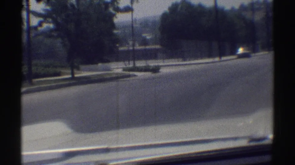 1969 Woodland Hills home-movie frame looking from a car window toward a quiet road, trees, curb, and fenced property beyond.