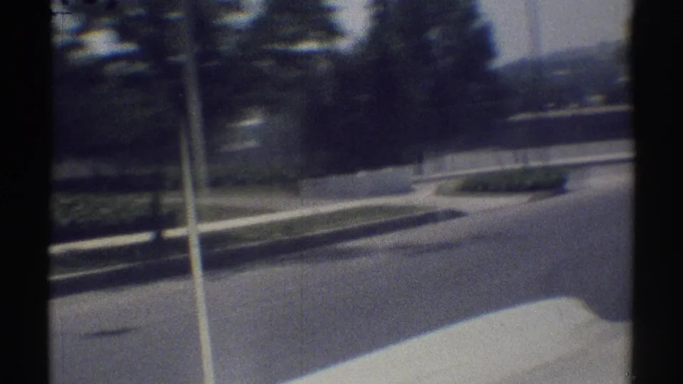 1969 Woodland Hills street scene from a moving car, with a roadside pole, curb, trees, and a fenced property in view.