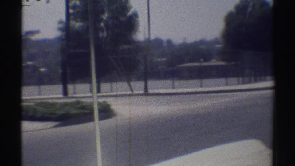 1969 Woodland Hills frame showing a curved suburban road, curb planting, utility poles, trees, and fenced grounds from a car.