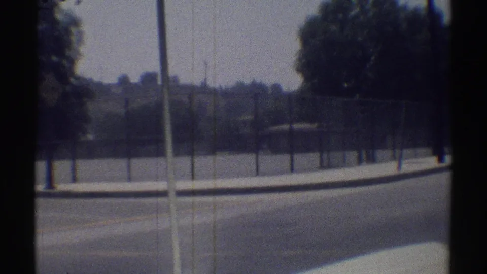 1969 Woodland Hills home-movie view of a fenced property, trees, curb, and road edge filmed from a passing car.