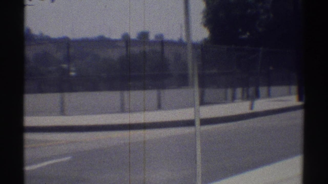 Suburban road beside a sidewalk and chain-link fence, seen from a moving vehicle.