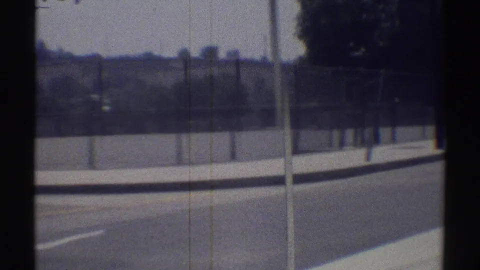 1969 Woodland Hills street view from a moving car, with pavement, curb, sidewalk, fence, trees, and soft 8mm blur.