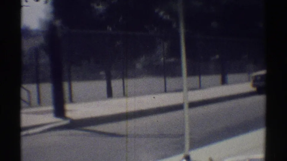 Woodland Hills roadside scene with chain-link fencing, trees, sidewalk, poles, and a partial car at frame edge on 8mm film.