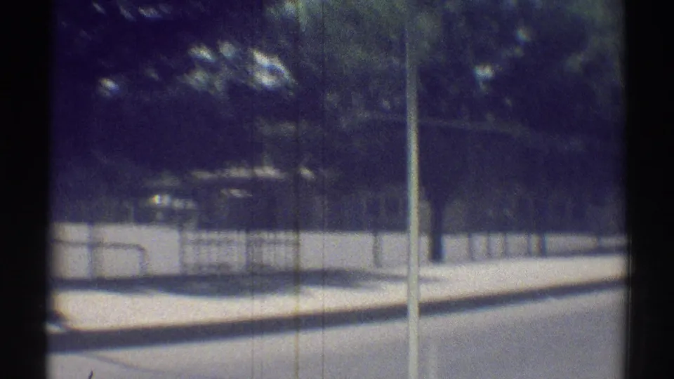 Moving 8mm view of a Woodland Hills roadside with chain-link fence, trees, sidewalk, curb, and quiet suburban street.