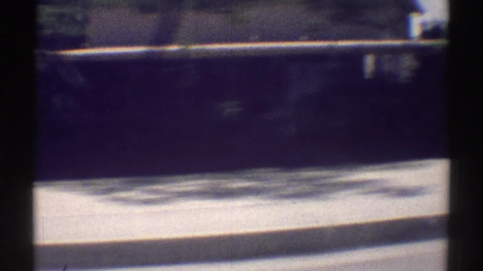 1969 Woodland Hills home-movie frame showing a moving car view past a bright curb, low wall, and dark tree shadows.