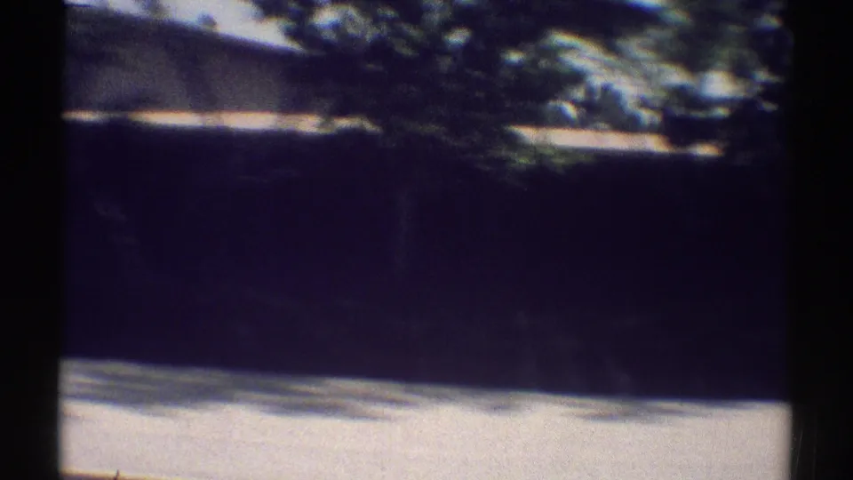 1969 8mm driving frame showing a blurred residential wall, roofline, trees, and sunlit sidewalk edge in Woodland Hills.