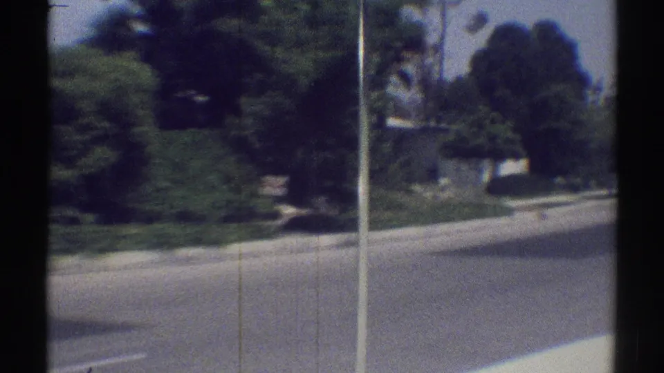 1969 Woodland Hills street view filmed from a moving car, with trees, curbside planting, and residential landscaping beyond the window frame.