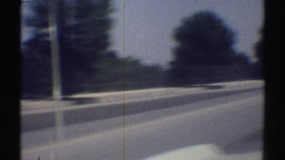 Woodland Hills road view from a moving car, with a curved road edge, trees, a pole, and dark interior shadows on grainy 8mm film.
