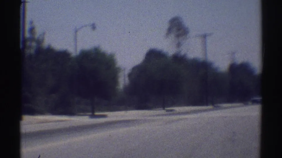 1969 Woodland Hills driving view with a curved road, roadside trees, light poles, and a distant vehicle softened by motion blur.