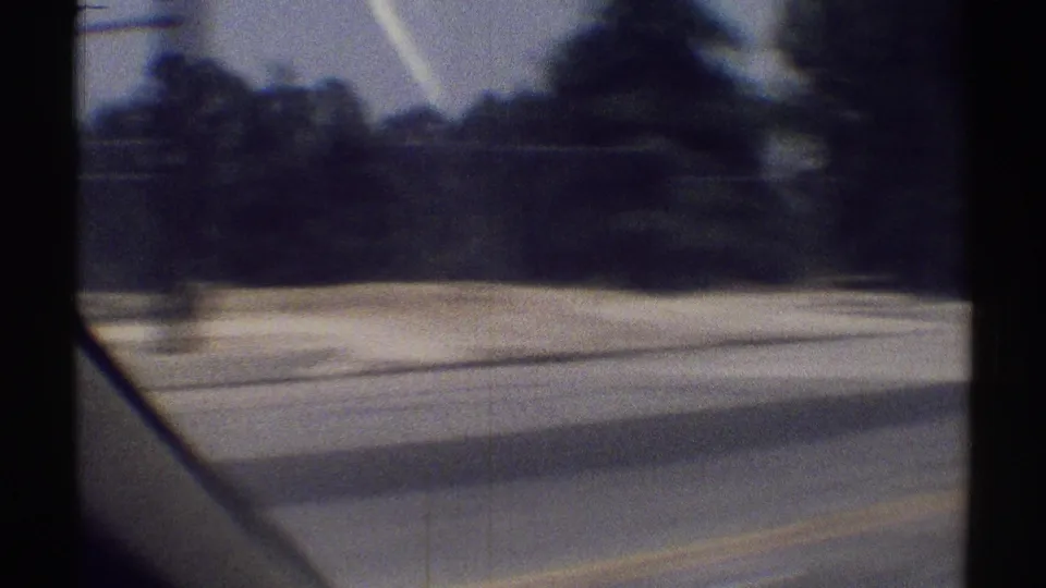 Woodland Hills driving frame from 1969 showing pavement, roadside trees, shadows, and part of the filming car or windshield edge.