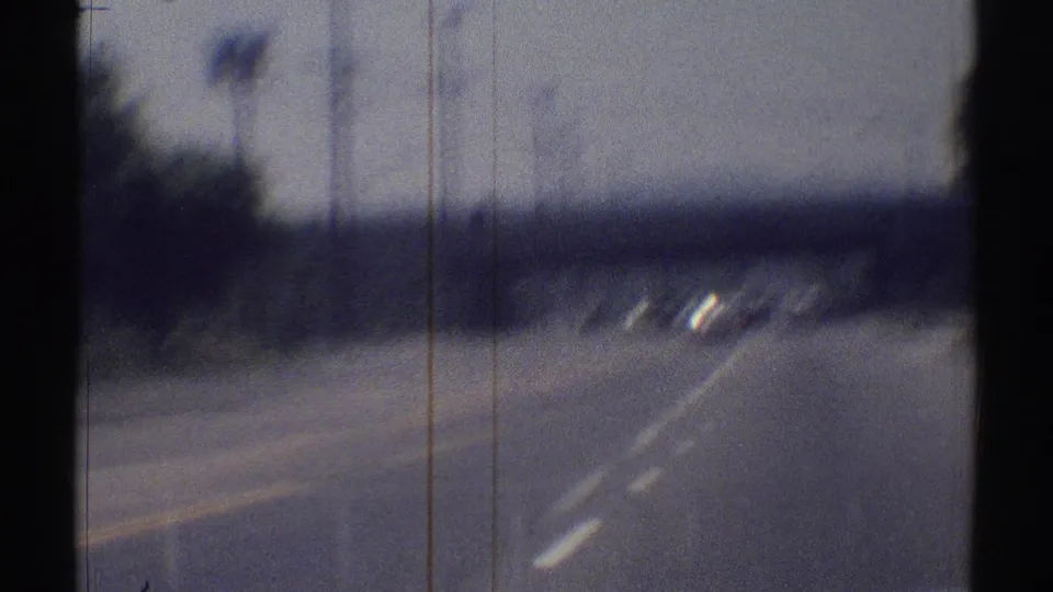 1969 Woodland Hills road view with lane markings, several cars ahead, roadside poles, trees, and low hills in the distance.