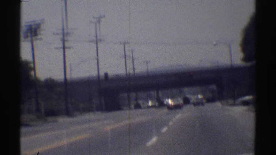 1969 Woodland Hills street view from moving 8mm footage, with cars ahead, lane markings, utility poles and an overpass.