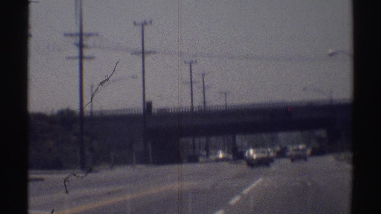Moving 8mm view of a Woodland Hills road in 1969, showing traffic under an overpass with poles and signals nearby.
