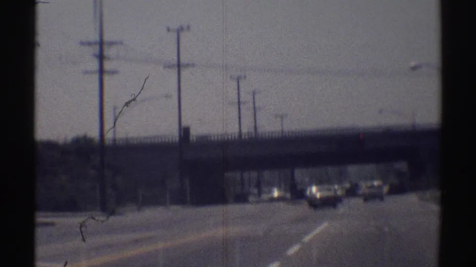 Moving 8mm view of a Woodland Hills road in 1969, showing traffic under an overpass with poles and signals nearby.
