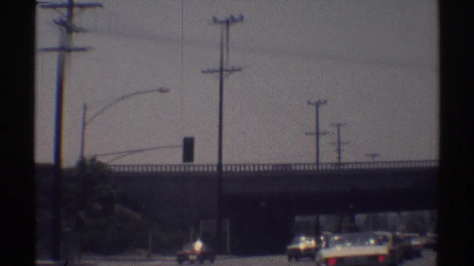 1969 Woodland Hills road scene with cars moving toward an overpass, utility poles, traffic signals, and soft 8mm blur.